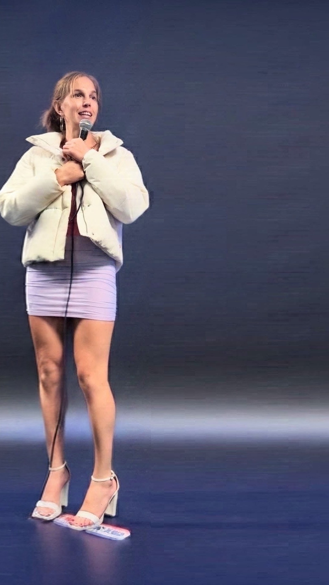 Nikki Brooklyn, stand-up comic, performing on stage under a spotlight, holding a microphone with both hands and wearing a cream puffer jacket